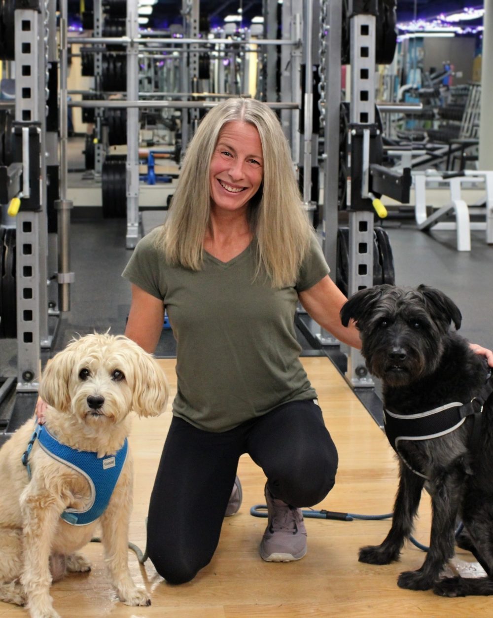 Tammy, group fitness and yoga instructor, kneeling with two dogs on the gym floor
